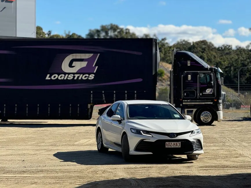 White sedan parked in front of a logistics truck at an outdoor loading area on a sunny day.